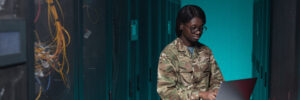 Portrait of young African-American woman wearing military uniform using computer while setting up network in server room, copy space