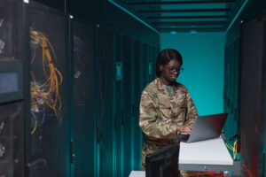 Portrait of young African-American woman wearing military uniform using computer while setting up network in server room, copy space